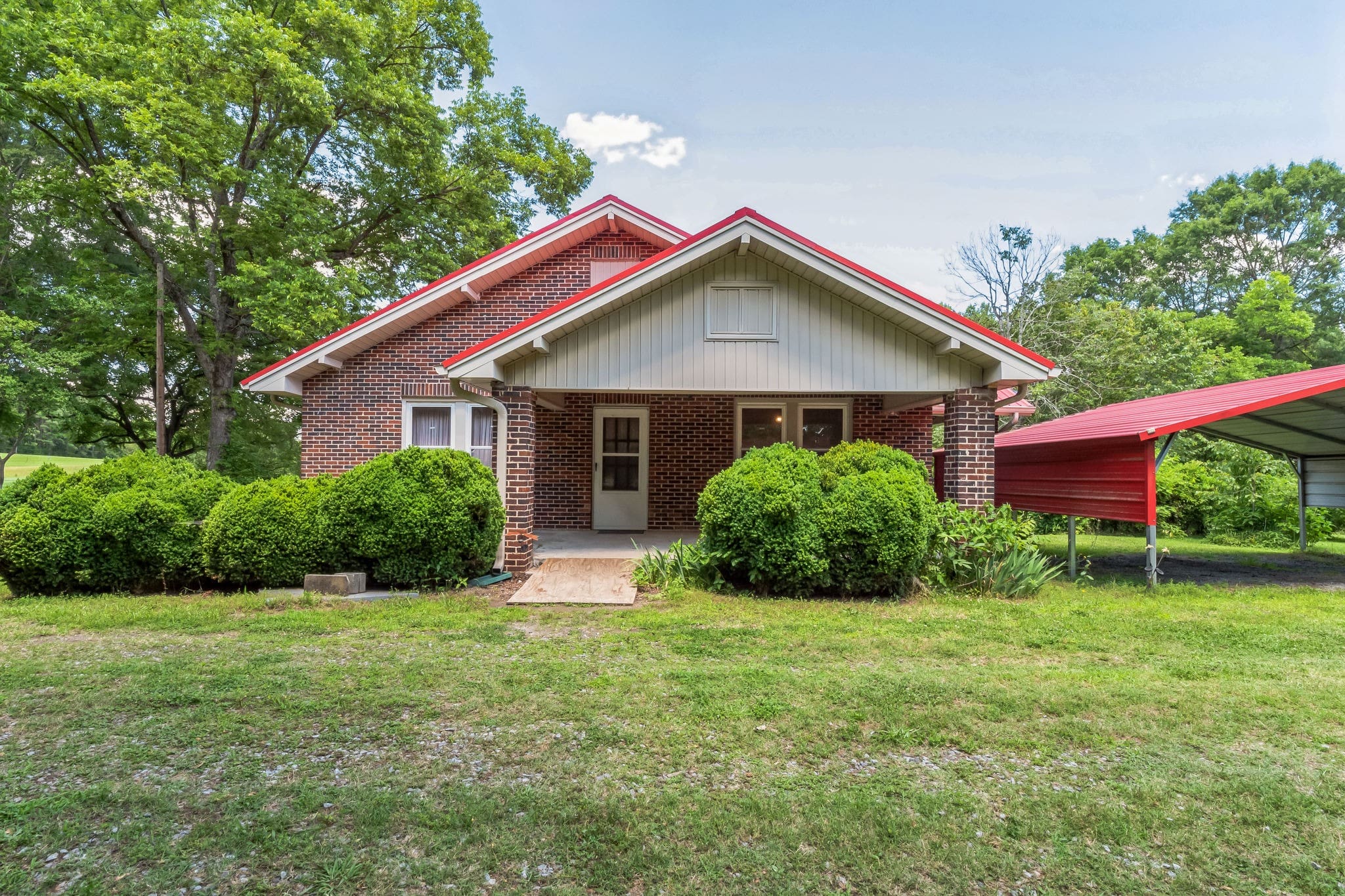 Charming brick house with a red roof, surrounded by lush greenery and neatly trimmed bushes, featuring a carport on the right side.