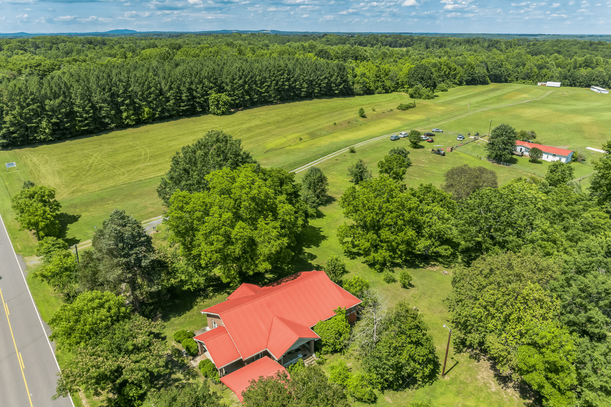 Aerial view of a rural property featuring a red-roofed house, lush greenery, and a nearby road with parked vehicles, showcasing a serene landscape.