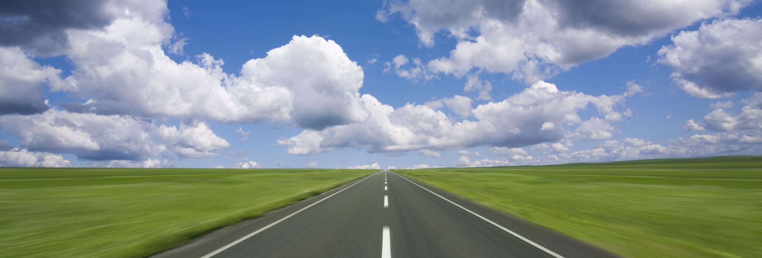 A long, straight road stretches into the distance, bordered by lush green fields under a bright blue sky filled with fluffy clouds.